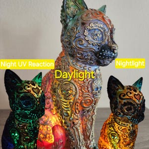 May include: Three decorative cat figurines with detailed, colourful designs. The central cat is illuminated, showing details under daylight. The left cat glows green under UV light, and the right cat glows orange in nightlight. The figurines feature floral and skull motifs.