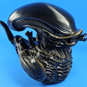 May include: A black and gold alien creature figurine with a detailed, textured surface. The creature has a large head with a dome-shaped top and a long, thin tail.
