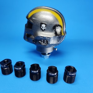 Op de afbeelding: Een zilver-geel helmvormig object met een schedel-embleem, gemonteerd op een metalen basis. Vijf zwarte schroefdraadadapters zijn eronder geplaatst. De achtergrond is effen blauw.