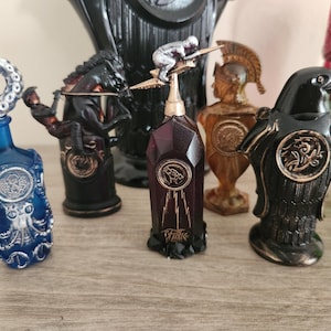 Op de afbeelding: Een verzameling decoratieve glazen flessen met unieke ontwerpen. De flessen hebben verschillende vormen en thema's, waaronder een octopus, een paard en ruiter, een figuur op een paal, een Romeinse soldaat en een raaf. De flessen zijn in tinten blauw, zwart, bruin en groen.