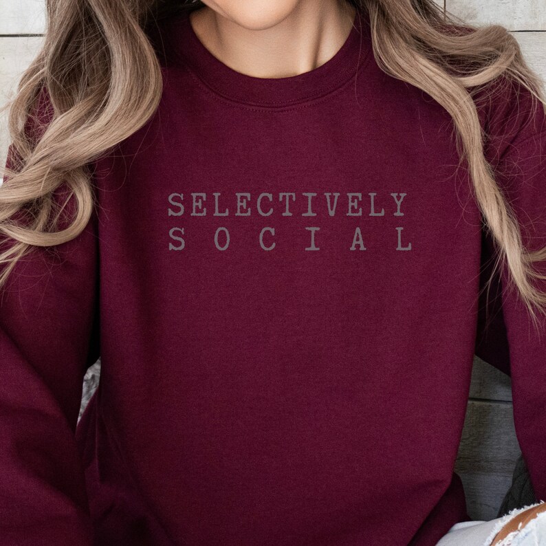 May include: A burgundy sweatshirt with the text "SELECTIVELY SOCIAL" in gray lettering. The crew neck sweatshirt is made of a soft material and is a solid color.