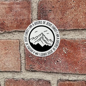 May include: A round white sticker with black text and graphics. The text reads "I AM A DISCIPLE OF JESUS CHRIST" around the border. Inside the circle is a mountain range and two birds. The sticker is on a brick wall.