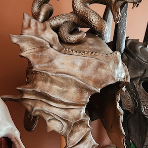 Könnte beinhalten: Ein goldener Helm mit einem geschnitzten Schlangenmotiv auf der Oberseite. Der Helm hat eine strukturierte, schuppige Oberfläche und ein spitzes, flügelartiges Design auf der Rückseite.