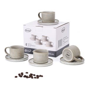 May include: A set of four espresso cups and saucers with a ribbed design. The cups and saucers are a neutral gray color. The box reads "4 Espresso Cups and Saucers". Coffee beans are scattered in front of the set.