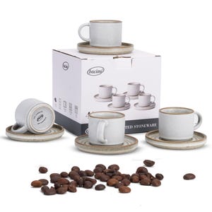 May include: A set of four white stoneware coffee cups and saucers with a light brown rim. The cups and saucers are arranged around a box with the brand name "bacimi" printed on it. Coffee beans are scattered in the foreground.