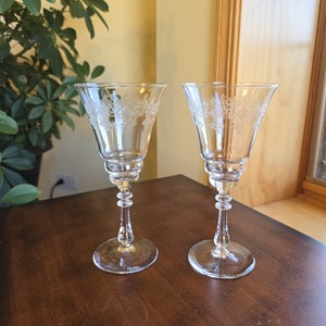 May include: Two clear, stemmed wine glasses with etched floral designs. The glasses have a flared bowl, a detailed stem, and a round base. They are displayed on a dark wooden surface, with a leafy plant and a window frame in the background.