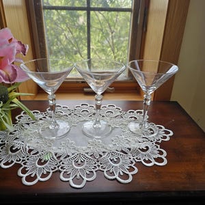 May include: Three clear glass martini glasses with wide, conical bowls and slender, multi-faceted stems. Each glass features a circular base and a transparent finish, arranged in a row on a white lace doily with intricate floral patterns.