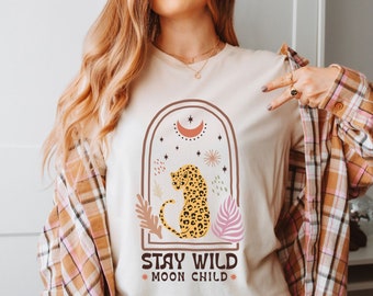 May include: A beige t-shirt with a graphic of a leopard sitting in a crescent moon arch with stars and floral accents. The text "Stay Wild Moon Child" is printed below the graphic.