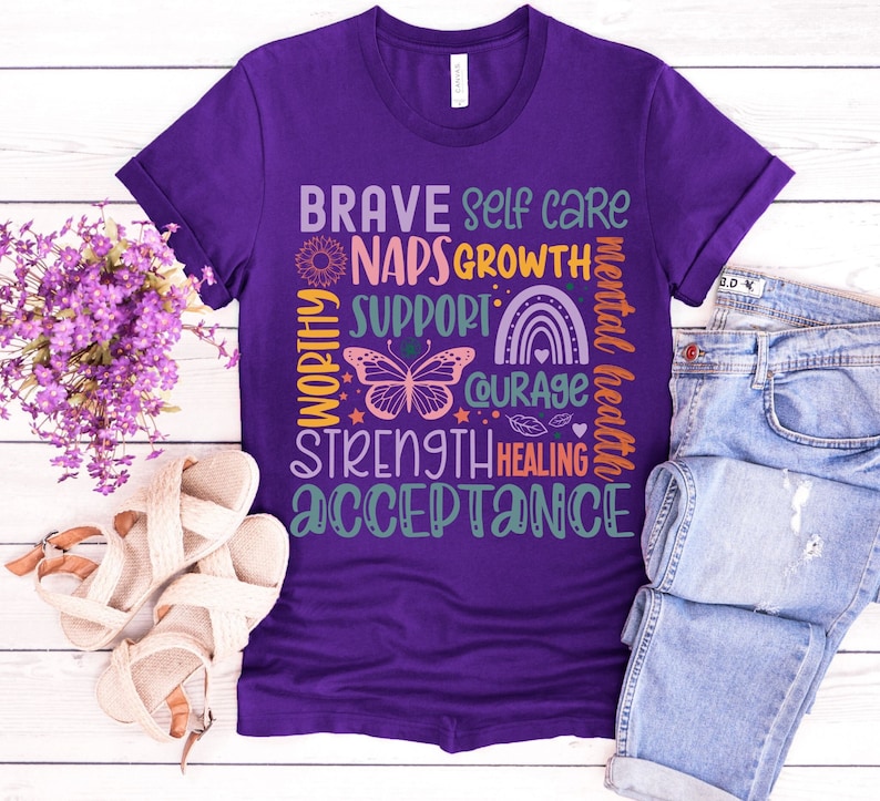 以下が含まれることがあります： 紫色のTシャツに、カラフルなグラフィックデザインが施されています。デザインには、「Brave」、「Self Care」、「Naps」、「Growth」、「Support」、「Courage」、「Mental Health」、「Strength」、「Healing」、「Acceptance」の言葉が書かれています。