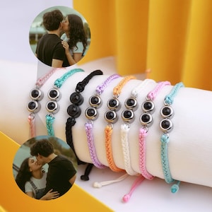 May include: A selection of friendship bracelets in various colours. Each bracelet has three black beads in silver-coloured settings, threaded on braided cords. The colours include pink, teal, black, purple, orange, and white. The bracelets are displayed on a white surface.