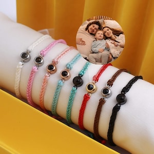 Puede incluir: Una colección de pulseras trenzadas coloridas con un dije circular. Las pulseras son de varios colores, incluyendo blanco, rosa, melocotón, turquesa, rojo, marrón y negro. Una pequeña foto de una familia está en el fondo.