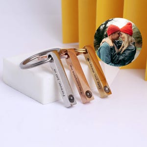 May include: Three metal keychains in silver, rose gold, and gold. Each keychain has a rectangular bar with engraved text: "Forever, just us", "You complete me", and "Only you, always". A small photo of a couple is in the background.