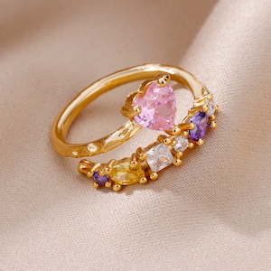 Peut inclure: Bague en or avec une pierre en forme de cœur rose et de plus petites pierres colorées. La bague est conçue pour être portée sur le majeur.