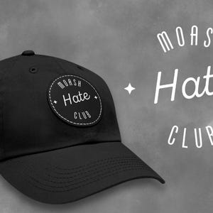 Può includere: Cappellino da baseball nero con una toppa nera e bianca che dice "Moash Hate Club".