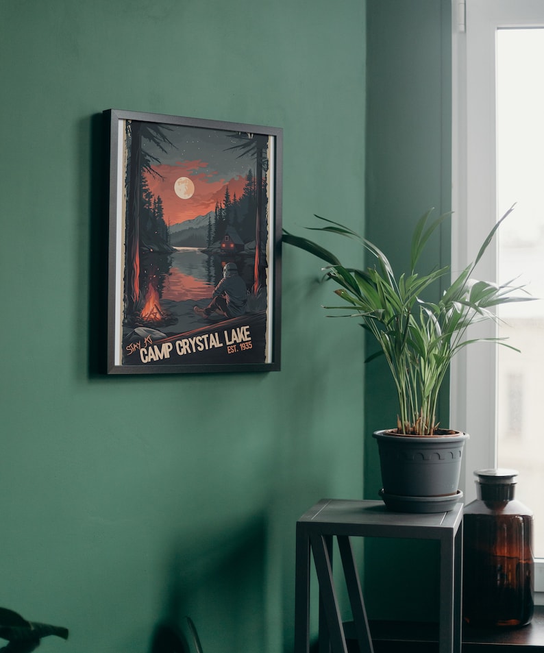 May include: A framed poster with a vintage aesthetic depicting a lake scene with a cabin, a campfire, and a full moon. The text on the poster reads "Stay at Camp Crystal Lake Est. 1955".