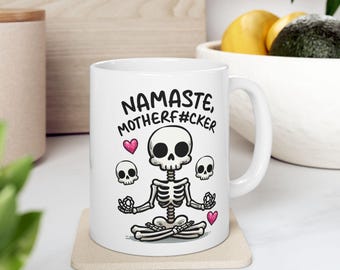 May include: White ceramic coffee mug with a black and white illustration of a skeleton doing yoga. The text "Namaste, Motherf#cker" is printed on the mug.