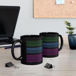 May include: Two black ceramic mugs with colorful geometric patterns. Each mug features a series of horizontal lines in green, blue, and pink. The mugs are on a light-colored wooden surface.