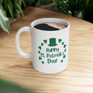 May include: White ceramic mug filled with dark liquid, featuring a green leprechaun hat and shamrocks around the text "Happy St. Patrick's Day!" on a wooden surface. The mug has a white handle.