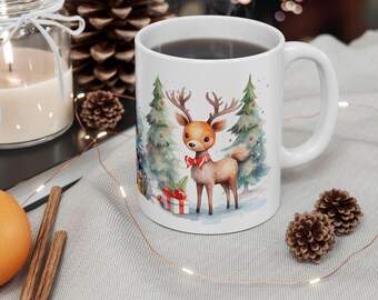 May include: White ceramic mug with a watercolor illustration of a deer in a winter forest setting. The deer is wearing a red bow and is standing near a Christmas tree and a gift box. The mug is filled with coffee.