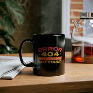 May include: Black ceramic mug with the text "ERROR 404 MOTIVATION NOT FOUND" in red and yellow. The mug is on a wooden surface, with steam rising from it. A glass carafe is in the background.