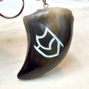 May include: A curved, claw-shaped keychain pendant made of polished, translucent dark brown and black horn material. A white, hand-painted geometric symbol is centered on the front surface. The pendant is attached to a silver-toned metal ring.