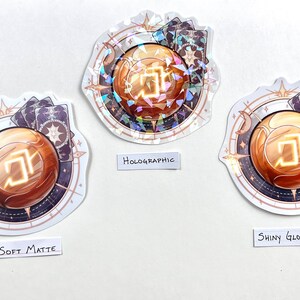 Peut inclure: Trois autocollants avec un motif d'orbe dor&eacute;, chacun avec une finition diff&eacute;rente : mat doux, holographique et brillant. L'orbe est entour&eacute; d'un motif de type compas avec des &eacute;toiles et des cartes.