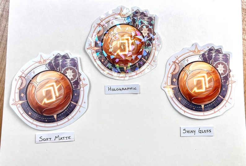 Peut inclure: Trois autocollants avec un motif d'orbe dor&eacute;, chacun avec une finition diff&eacute;rente : mat doux, holographique et brillant. L'orbe est entour&eacute; d'un motif de type compas avec des &eacute;toiles et des cartes.