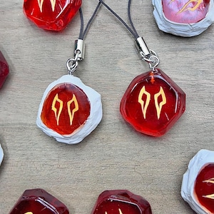 May include: Close-up of several red and white pendants with gold-colored symbols. Each pendant has a unique shape and is attached to a black cord with a silver clasp. The pendants are arranged on a wooden surface.