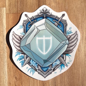 May include: A white sticker with a blue crystal in the center, surrounded by silver wings, swords, and floral designs. The crystal has a white shield with a cross inside. The sticker has the text "akumacon" at the bottom.