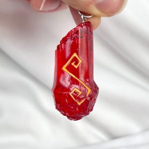 May include: A translucent red crystal pendant with a gold-colored symbol. The pendant is suspended from a silver-colored chain. The crystal has a faceted surface and is held up by a hand.