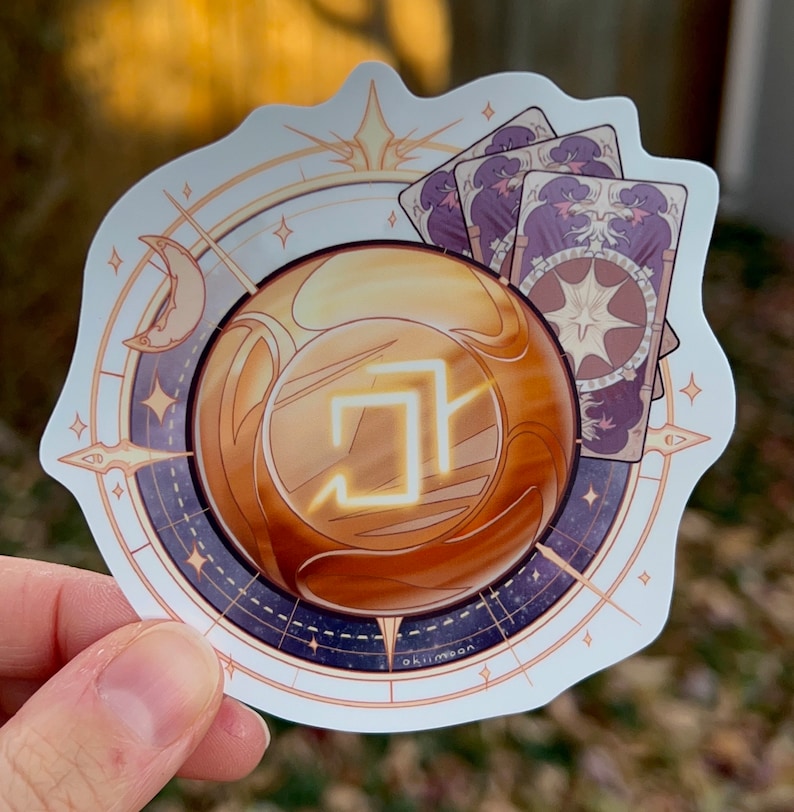 Peut inclure: Un autocollant blanc avec un orbe dor&eacute;, une boussole et trois cartes de tarot violettes. L'orbe contient un carr&eacute; lumineux. La boussole a un croissant de lune et une &eacute;toile. Les cartes de tarot repr&eacute;sentent une &eacute;toile et un croissant de lune. L'autocollant est marqu&eacute; "okimoon" en bas.