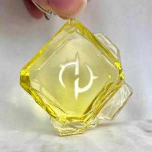 May include: A yellow, faceted keychain pendant with a white, stylized symbol. The pendant is held by a silver-colored keyring. The pendant has a geometric shape with a diamond-like center and additional facets.
