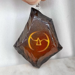 May include: A dark brown pendant with a silver-colored loop. The pendant is a rough, geometric shape with a glowing orange center. A yellow symbol is visible in the center of the pendant.