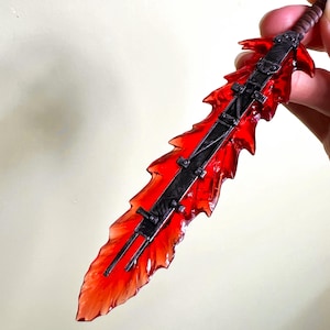 Puede incluir: Una espada de fantasía en miniatura con una hoja roja translúcida y detalles metálicos negros. La hoja tiene un diseño en forma de llama y la empuñadura es marrón. La espada se sostiene sobre un fondo claro.