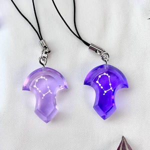 May include: Two purple resin pendants with a constellation design, each attached to a black cord with a silver clasp. The pendants have a translucent appearance and a geometric shape, with a white constellation pattern.