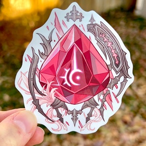 May include: A red gemstone with a stylized symbol inside, surrounded by intricate black and white designs. The design includes a cross, a sword, and a flower.