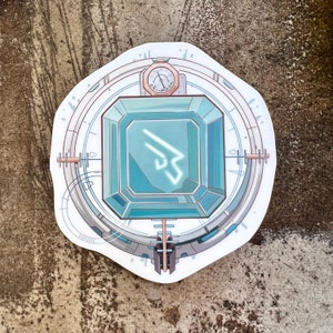 May include: A white sticker with a blue and green gem in the center. The gem has a white symbol inside. The sticker has a circular design with a clock face at the top.