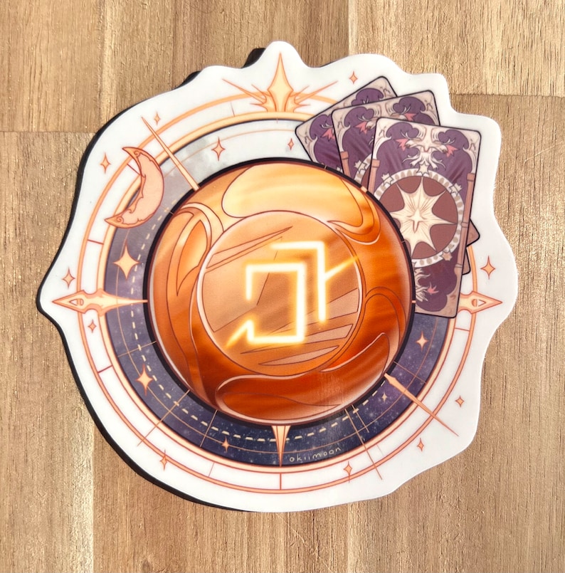 Peut inclure: Un autocollant repr&eacute;sentant un orbe dor&eacute; avec un symbole lumineux &agrave; l'int&eacute;rieur, entour&eacute; d'une boussole avec des &eacute;toiles et une lune croissante. Trois cartes &agrave; jouer aux motifs complexes sont visibles en arri&egrave;re-plan.