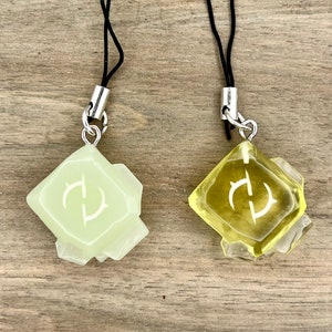 May include: Two glow-in-the-dark charm pendants with a black string. The charms are shaped like a crystal with a white symbol in the center. One charm is green and the other is yellow.