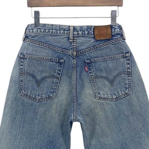 May include: A pair of blue denim jeans with a brown leather patch on the back. The jeans have a faded look and are hanging on a wooden hanger.