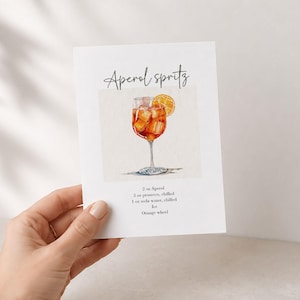 Pode incluir: Um cart&atilde;o retangular branco com uma ilustra&ccedil;&atilde;o em aguarela de um Aperol Spritz num copo com haste e uma fatia de laranja. O texto diz Aperol spritz, 2 oz Aperol, 3 oz prosecco, chilled, 1 oz soda water, chilled, ice, e Orange wheel.