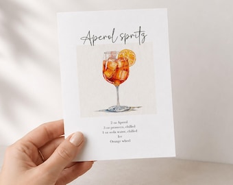 May include: A white rectangular card featuring a watercolor illustration of an Aperol Spritz in a stemmed glass with an orange slice. The text reads Aperol spritz, 2 oz Aperol, 3 oz prosecco, chilled, 1 oz soda water, chilled, ice, and Orange wheel.