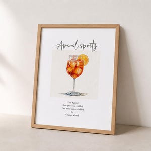 Pode incluir: Uma impress&atilde;o emoldurada com uma ilustra&ccedil;&atilde;o em aguarela de um Aperol Spritz num copo com uma rodela de laranja. O texto diz Aperol Spritz e lista os ingredientes: 2 oz Aperol, 3 oz prosecco, 1 oz &aacute;gua com g&aacute;s, gelo, rodela de laranja.