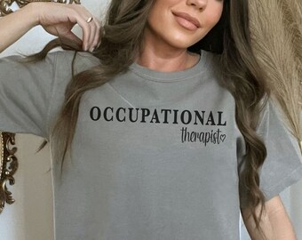 May include: A gray t-shirt with the words "OCCUPATIONAL therapist" printed in black. The shirt is a crew neck style and appears to be made of a soft material. The word "therapist" has a heart symbol next to it.