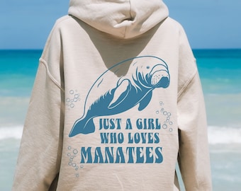 May include: Beige hoodie with a blue manatee graphic and the text "JUST A GIRL WHO LOVES MANATEES." The design features a detailed illustration of a manatee with bubbles. The hoodie is a casual wear item.