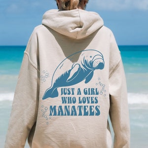 May include: Beige hoodie with a blue manatee graphic and the text "JUST A GIRL WHO LOVES MANATEES." The design features a detailed illustration of a manatee with bubbles. The hoodie is a casual wear item.