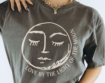 May include: A charcoal grey t-shirt with a graphic of a stylized moon face with closed eyes. The text "LOVE BY THE LIGHT OF THE MOON" is printed around the moon graphic.