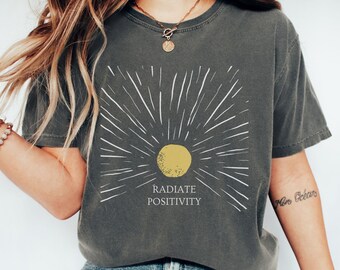 May include: A dark gray t-shirt with a white sunburst design and the text "RADIATE POSITIVITY" in white. The sun is yellow.