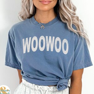 May include: A blue t-shirt with the word "WOOOWOO" printed in white on the front. The shirt is tied at the waist and the model is wearing it with grey sweatpants.