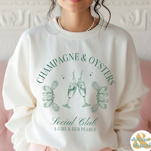 May include: White sweatshirt with a green graphic design that says "Champagne & Oysters Social Club A Girl & Her Pearls". The graphic features two champagne glasses clinking and oysters.